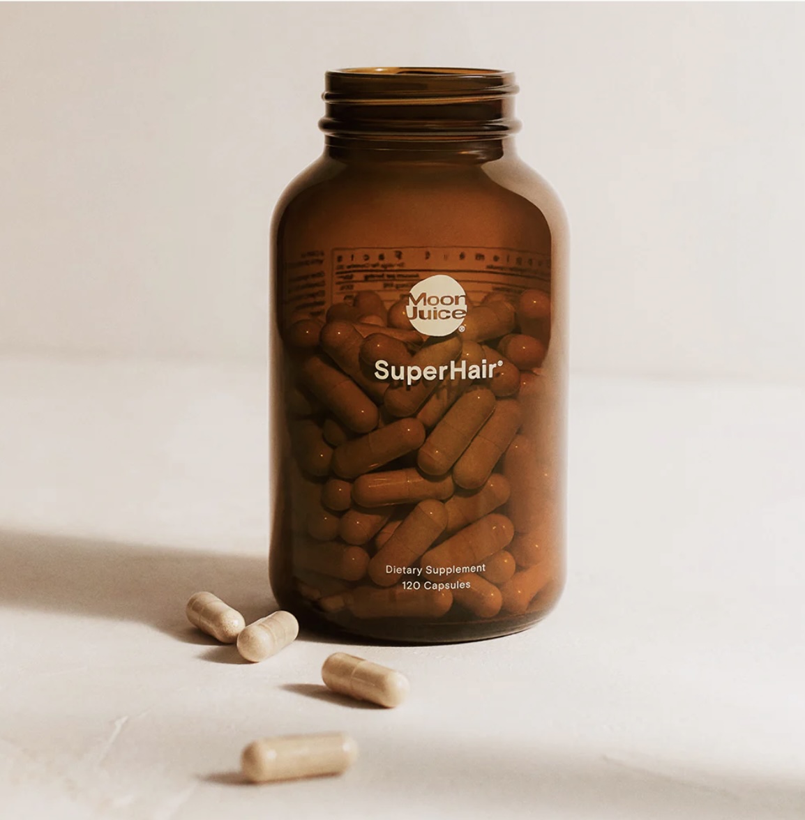 A brown glass bottle labeled "Moon Juice SuperHair" contains capsules, with three capsules lying on the white surface in front of it.