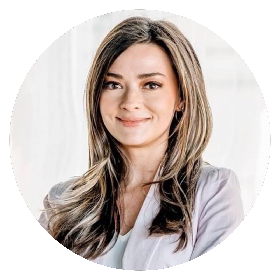 A woman with long, light brown hair and a light purple blazer smiles slightly while standing in front of a bright, blurred background.