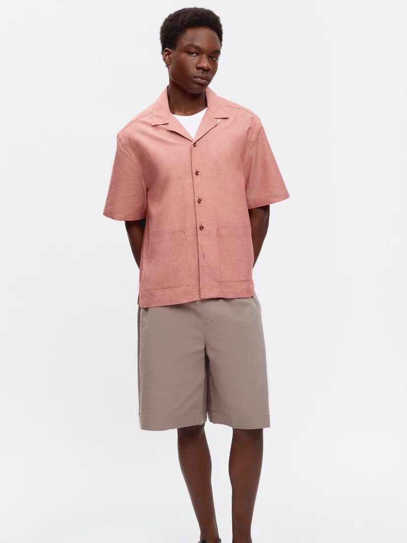 A man stands against a plain background wearing a pink short-sleeve button-up shirt, taupe shorts, black sandals, and a white undershirt.