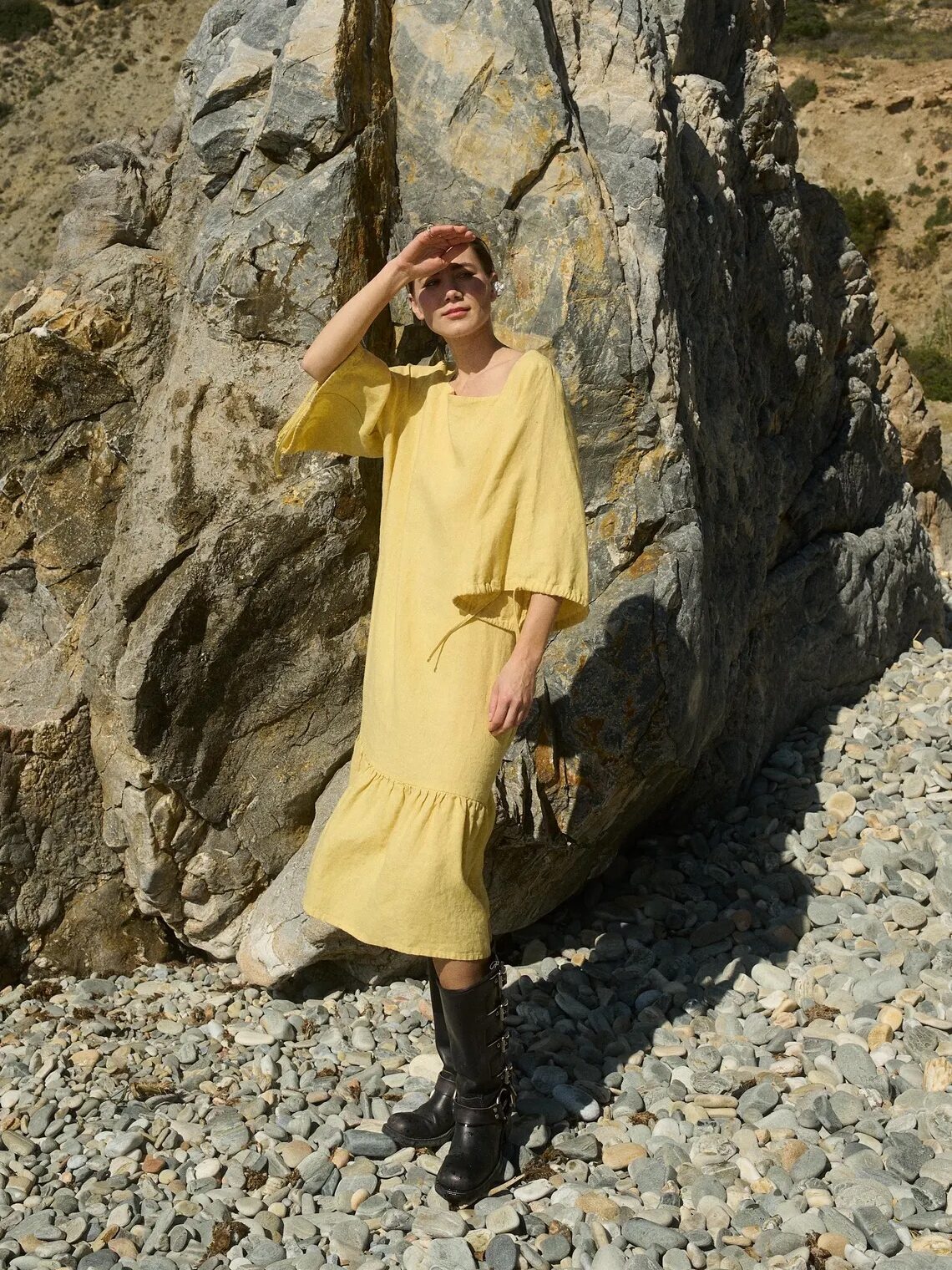 A person in a yellow dress and black boots stands on rocky ground, shielding their eyes from the sun, with large rock formations in the background.