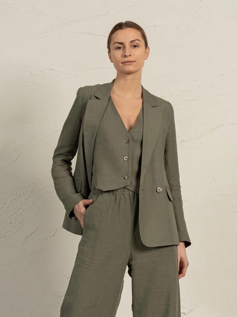 Woman wearing a light olive green tailored blazer over a matching buttoned vest and wide-leg trousers, standing against a textured off-white wall.