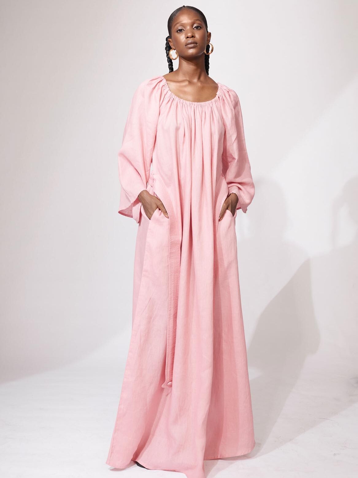 A person stands in a studio wearing a loose-fitting, long pink dress with long sleeves and pockets, looking directly at the camera.