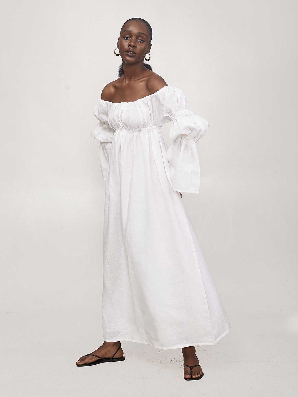 A woman wearing a white, off-the-shoulder, long-sleeve dress and black sandals stands against a plain light background.