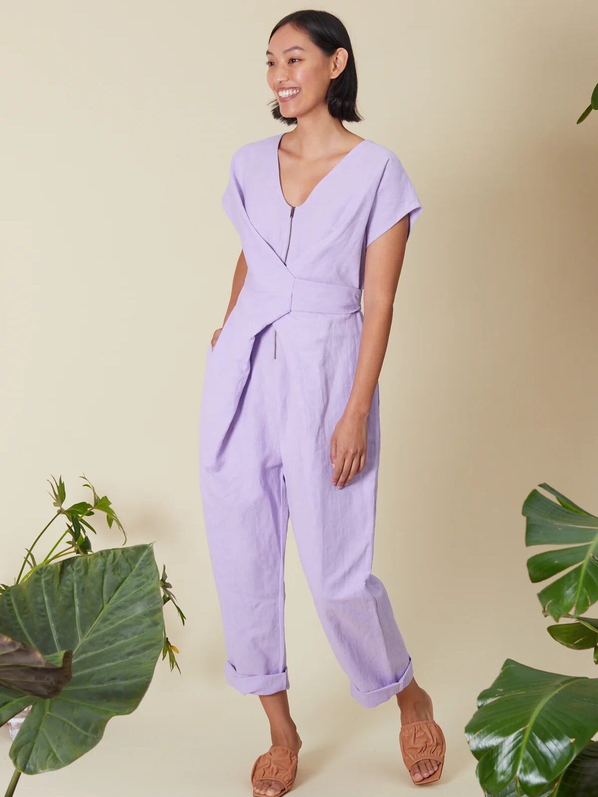 A woman stands smiling in a light purple jumpsuit with short sleeves and rolled-up pant legs, wearing tan sandals, surrounded by green plants against a beige background.