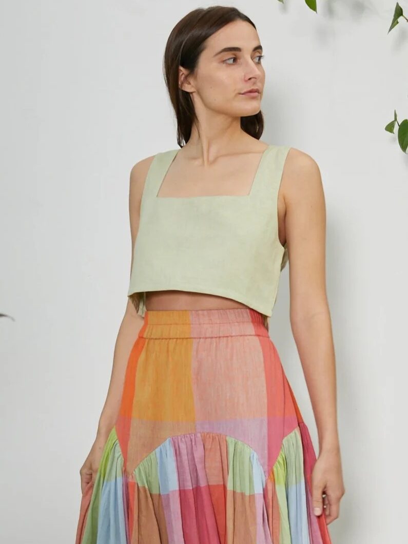 A woman wearing a light green square-neck crop top and a colorful, plaid tiered skirt stands against a plain background with some green leaves visible.