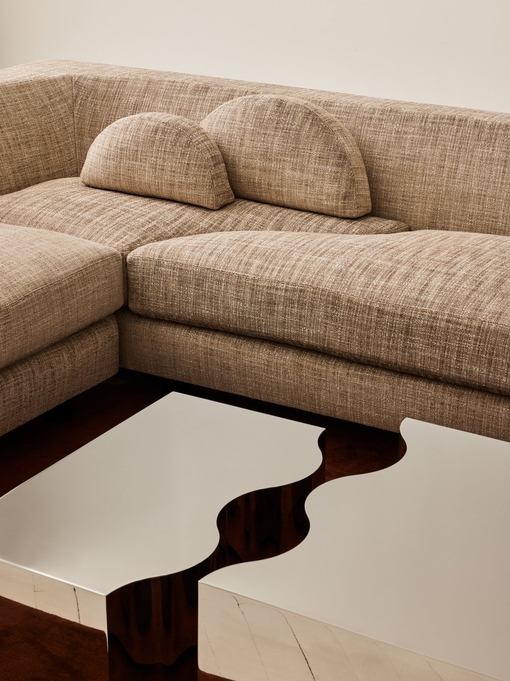 Beige textured sectional sofa with two semi-circular cushions, next to two metallic, wavy-edged coffee tables on a dark floor.
