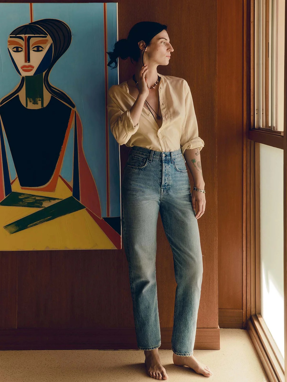 A barefoot woman in a beige shirt and jeans stands by a window, looking outside, with a stylized portrait painting on the wall behind her.