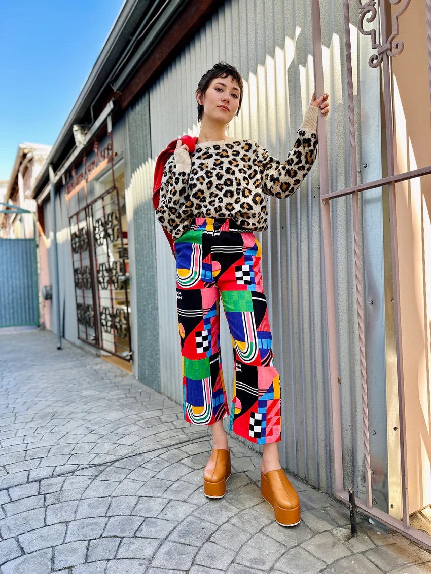 A person stands outdoors wearing a leopard print sweater, colorful geometric pants, tan platform shoes, and holding a red bag, beside a corrugated metal wall and gate.
