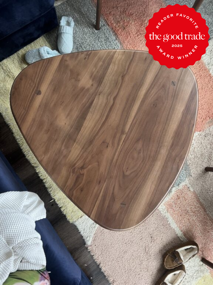 A wooden, triangular coffee table is placed on a multi-colored rug with a blanket, slippers, and part of a blue couch visible. A “Reader Favorite The Good Trade 2025 Award Winner” badge is in the corner.