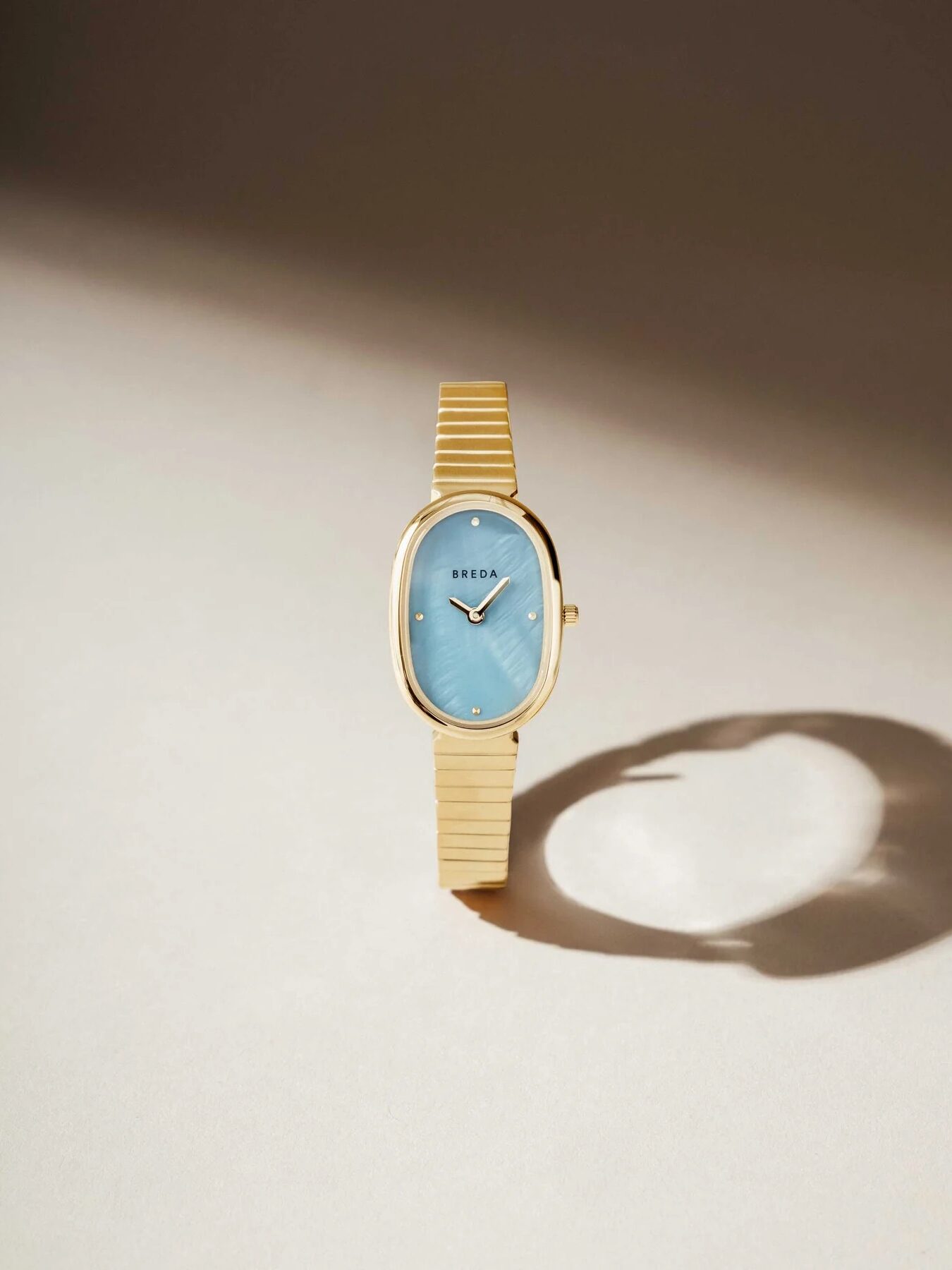 A gold wristwatch with a blue oval face and a minimalist design is placed on a light surface, casting a soft shadow.