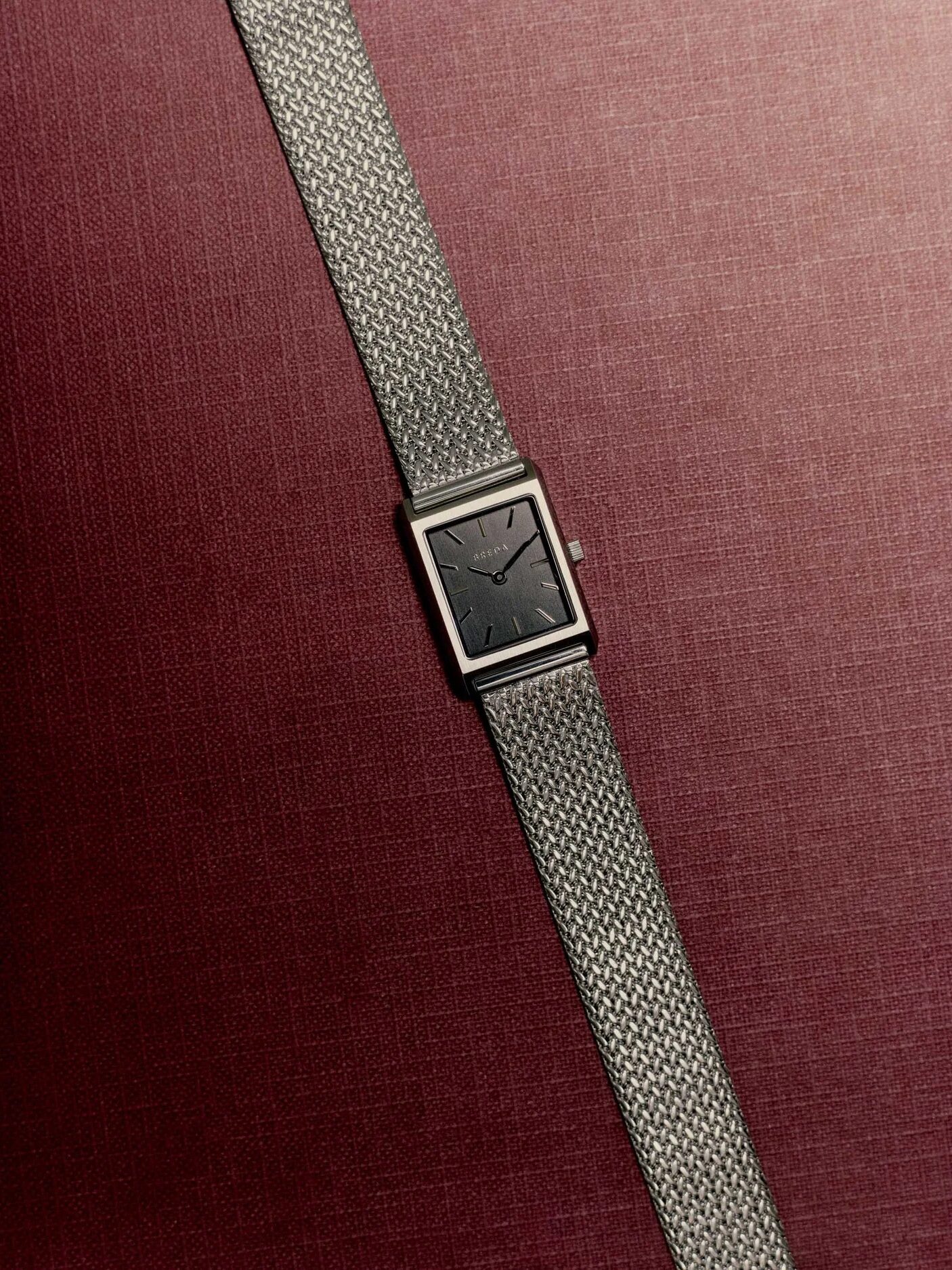 A rectangular silver wristwatch with a mesh band is displayed diagonally on a textured burgundy background.