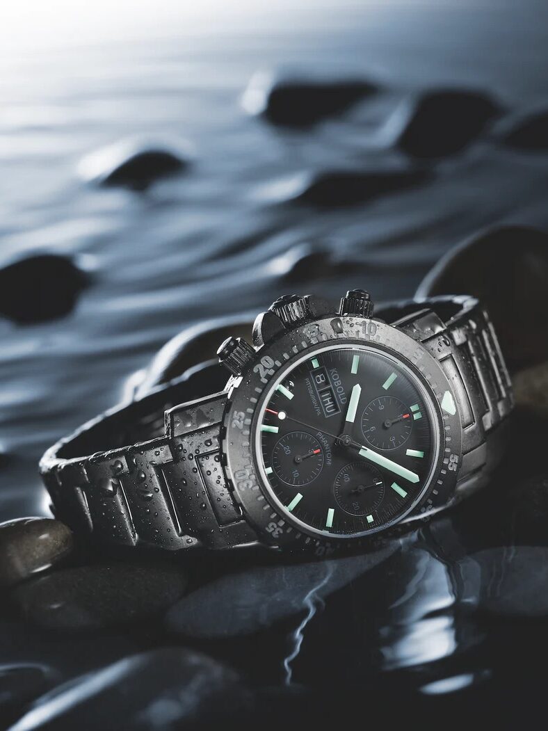 A stainless steel wristwatch with a black dial and glowing hands rests on wet rocks by the water.
