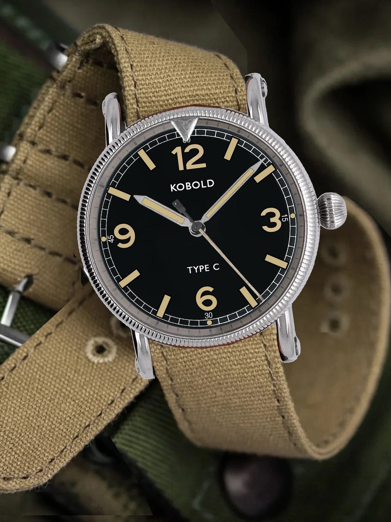 A Kobold Type C wristwatch with a tan fabric strap rests on green fabric, showing the time as 10:09:36. The watch has a black dial with beige numerals and hands.