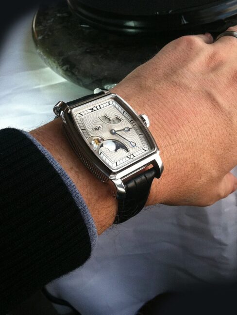 A person's left hand wearing a rectangular silver wristwatch with a black leather strap and a moonphase display, next to a marble surface.