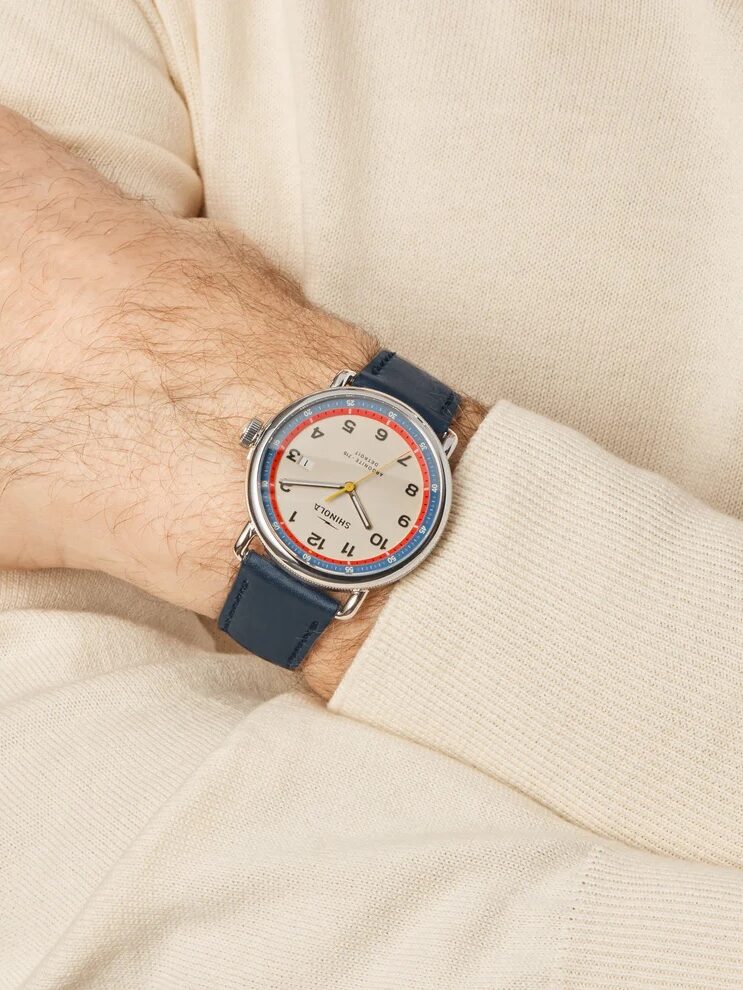 A person wearing a beige long-sleeve top displays a wristwatch with a cream dial, red and black numerals, and a blue leather strap.