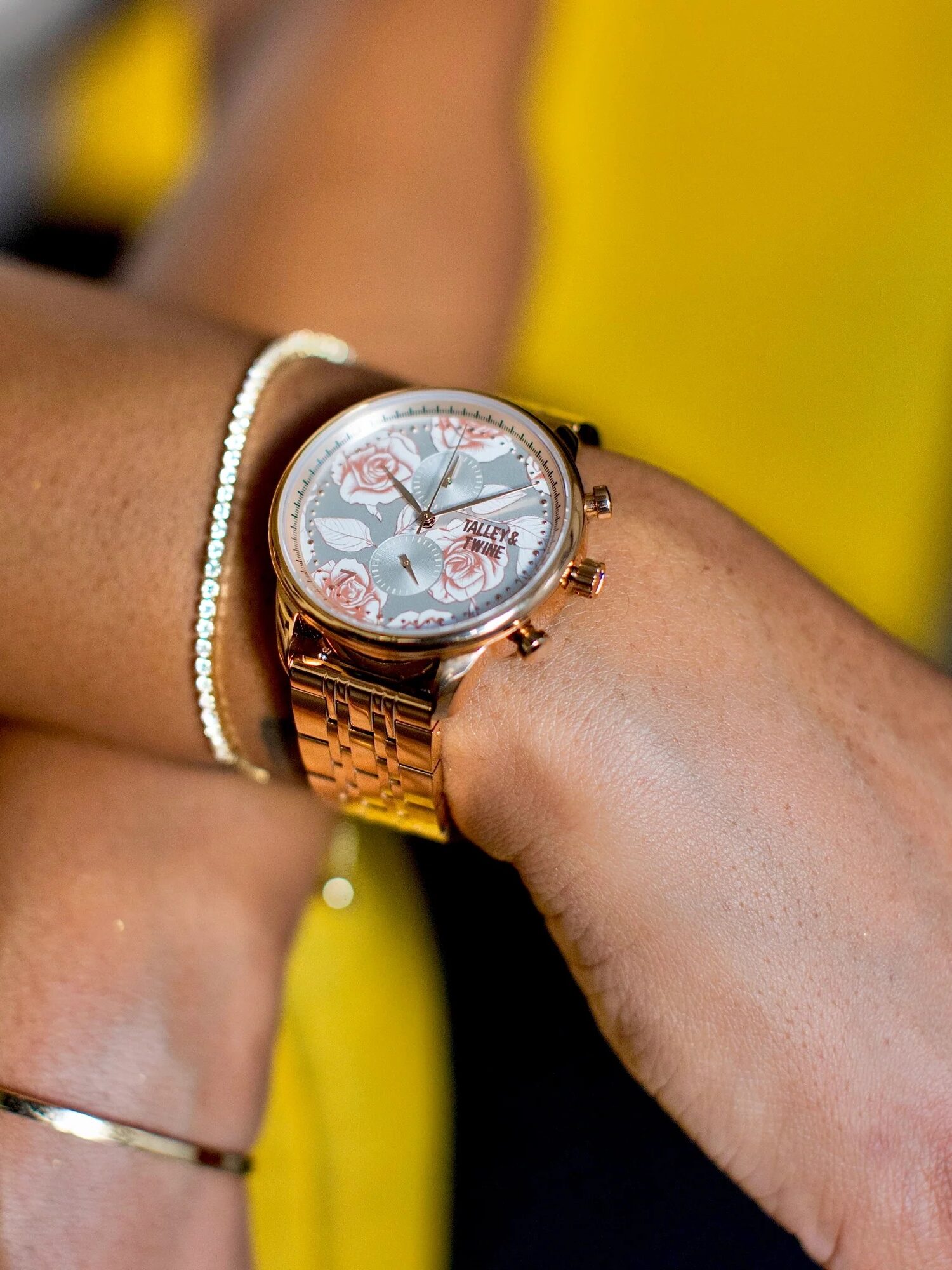A person wearing a gold wristwatch with a floral design on the face, along with two gold bracelets, against a yellow background.