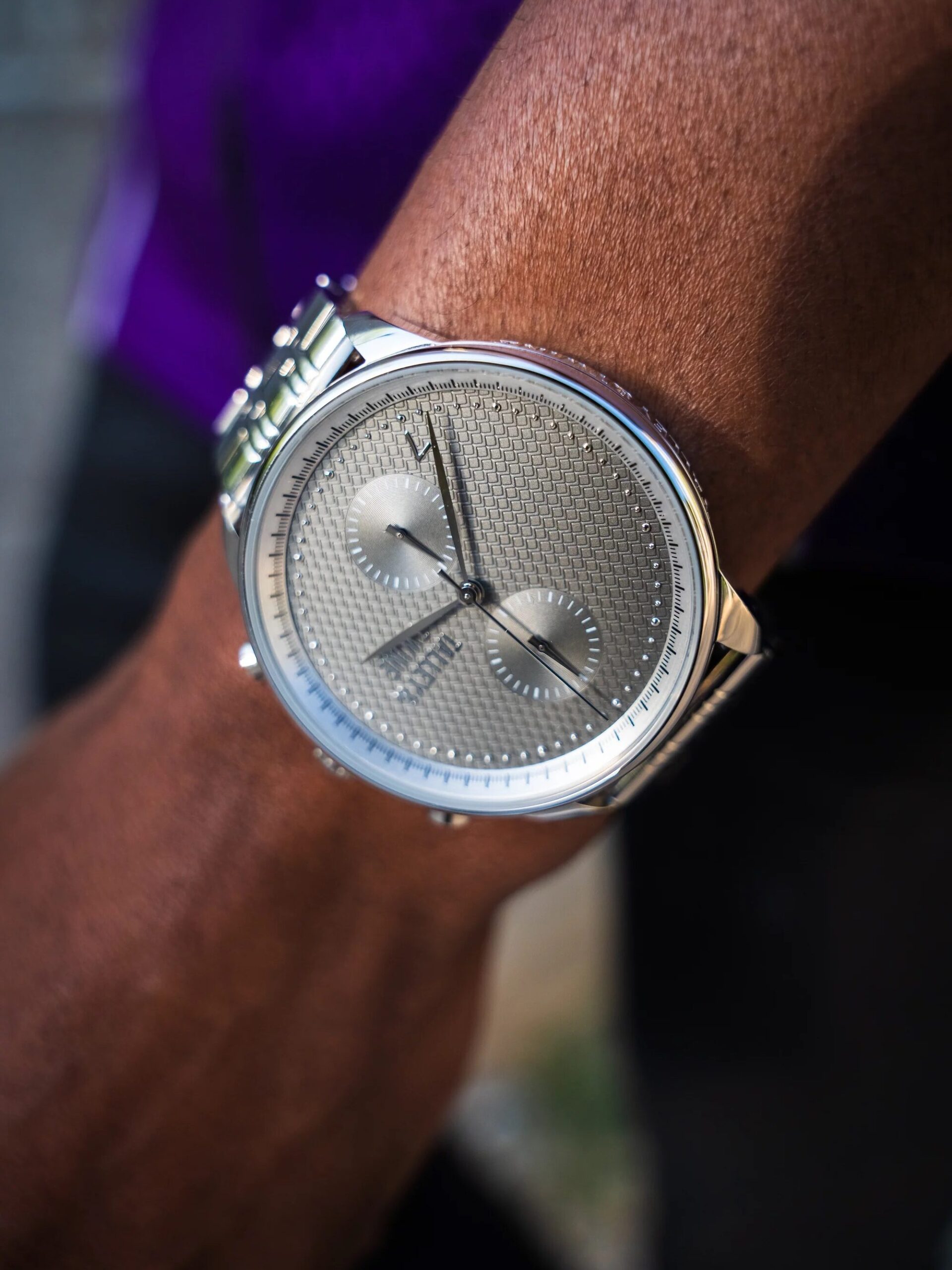 Close-up of a silver wristwatch with a textured face and two subdials on a person's wrist. The person is wearing a dark shirt.