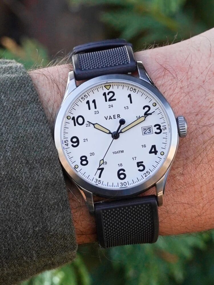 A wrist with a black-banded analog VAER watch displaying the time as 3:15:43 and the date as 23. The wearer has a green shirt and is outdoors.