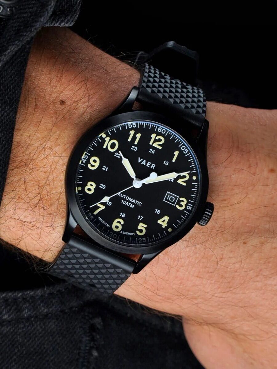 A person wearing a black wristwatch with a black strap, white numerals, and luminous hands; the watch displays the date and has a textured band.