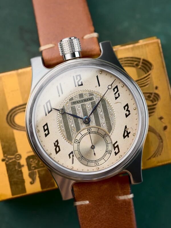 A wristwatch with a brown leather strap and a silver case, featuring black numerals and a small seconds subdial, rests on a gold-colored box with black graphics.