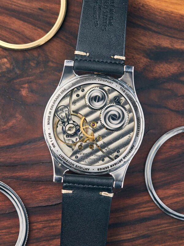 A wristwatch with a visible mechanical movement and black leather strap is placed on a wooden surface, surrounded by metal bangles.