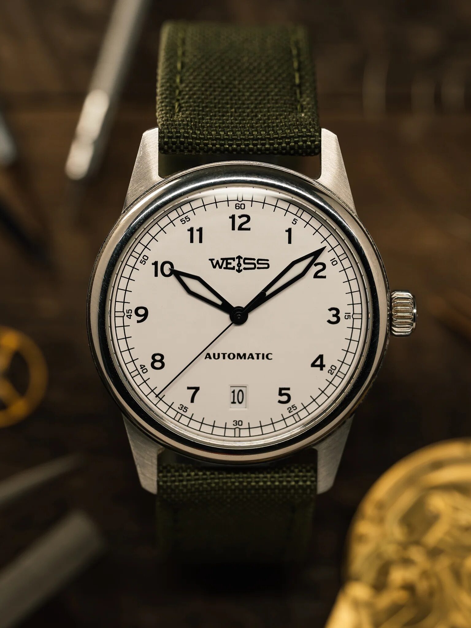 A Weiss automatic wristwatch with a white dial, black numerals, and an olive green strap is shown on a wooden surface with watchmaking tools nearby.