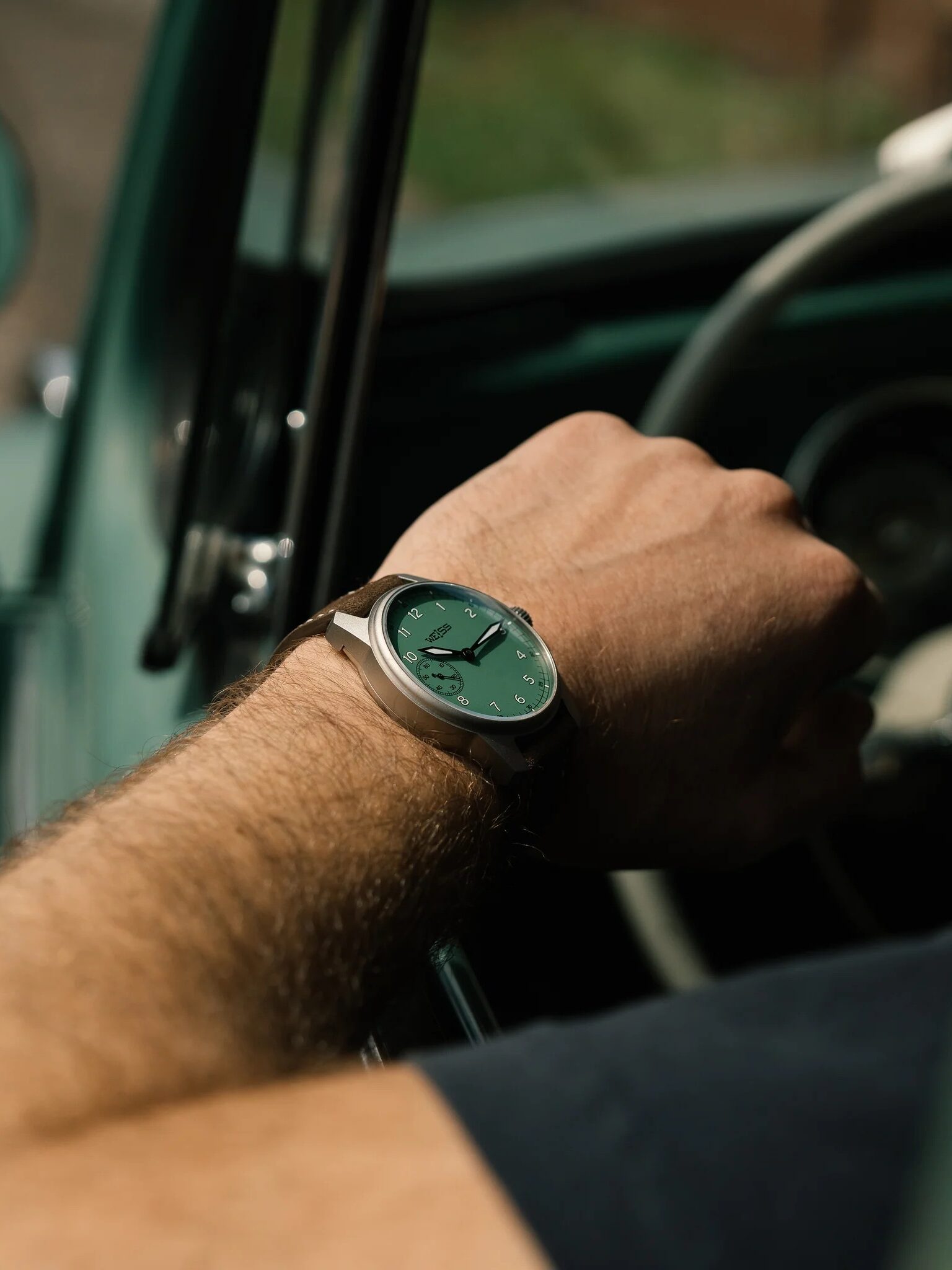A person wearing a green-faced wristwatch sits in the driver’s seat of a classic car with a green interior.