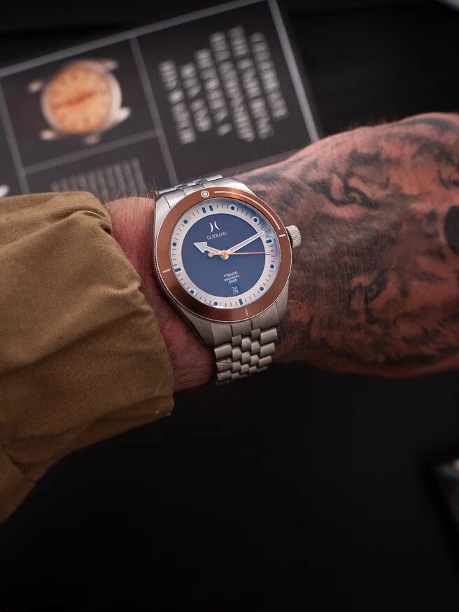 A person with a tattooed hand wearing a silver wristwatch with a blue and copper bezel dial, dressed in a tan sleeve.