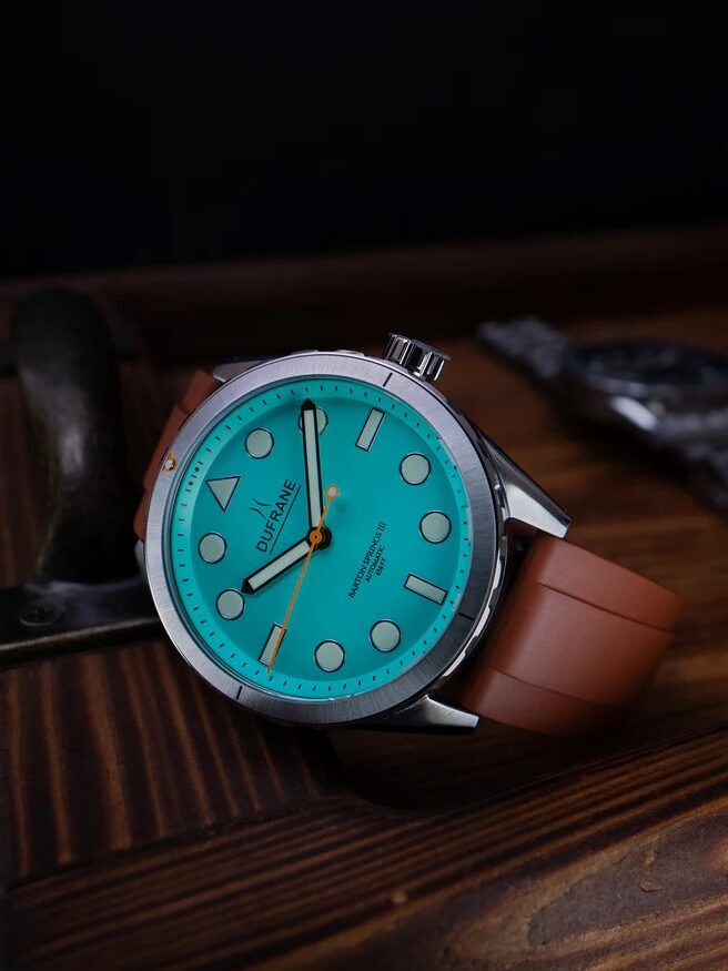 A wristwatch with a turquoise dial, silver markers, and a brown strap rests on a wooden surface next to a metal handle.