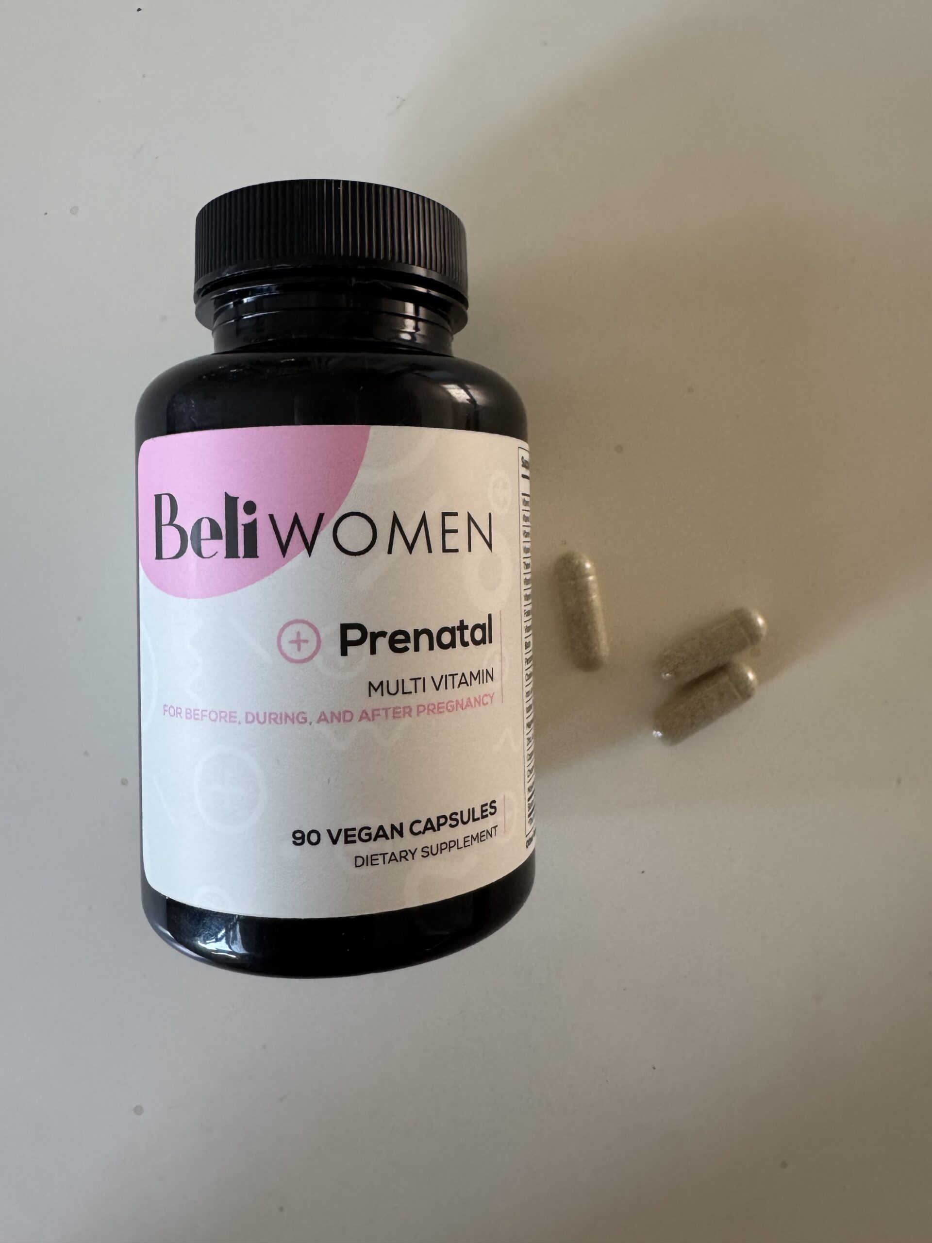 A bottle labeled "Beli Women Prenatal Multivitamin" with 90 vegan prenatal vitamin capsules sits next to two loose capsules on a white surface.