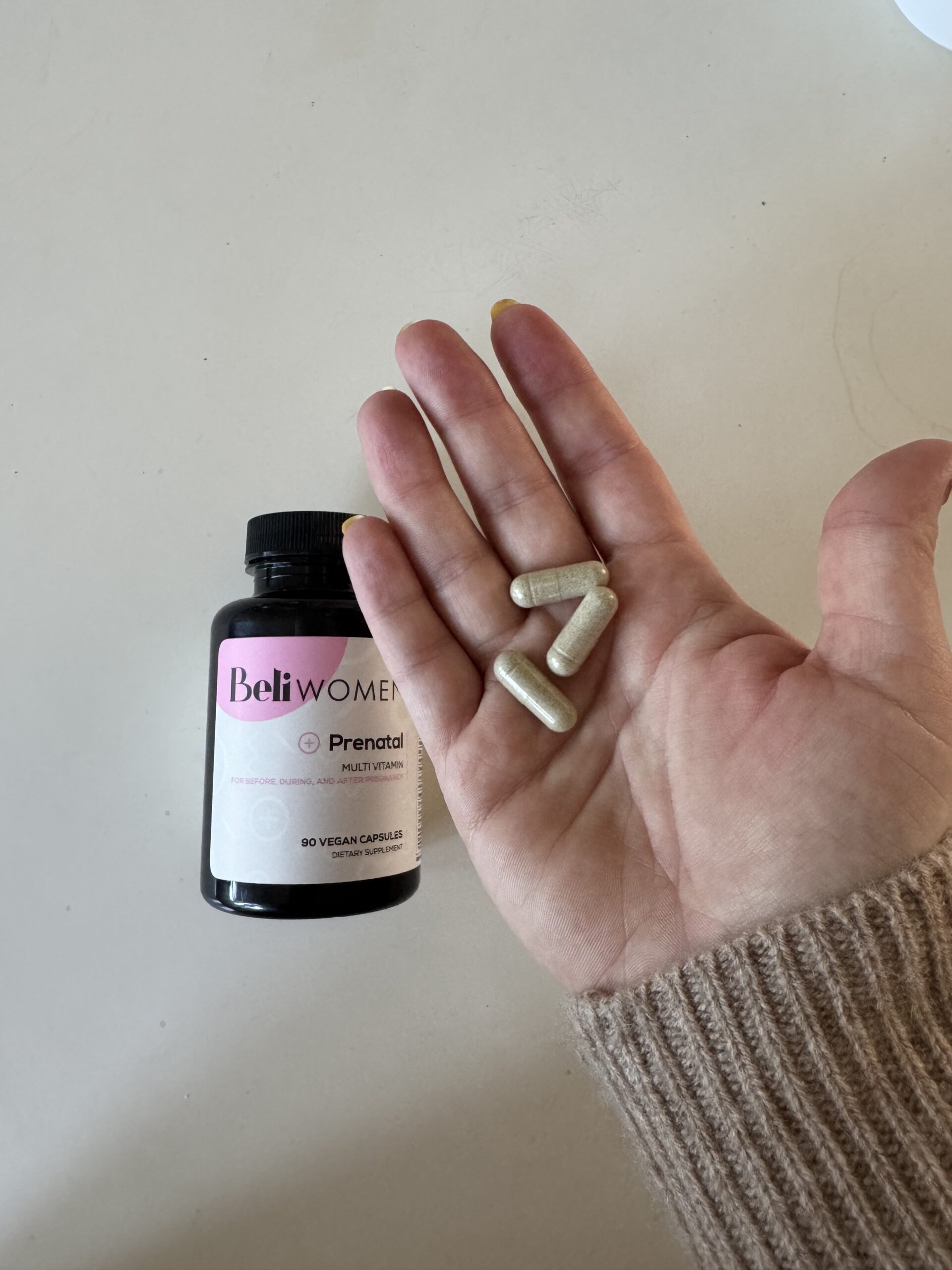 A hand holds three capsules next to a bottle labeled "Beli Women Prenatal Multivitamin," highlighting this prenatal vitamin as an essential supplement for expectant mothers.