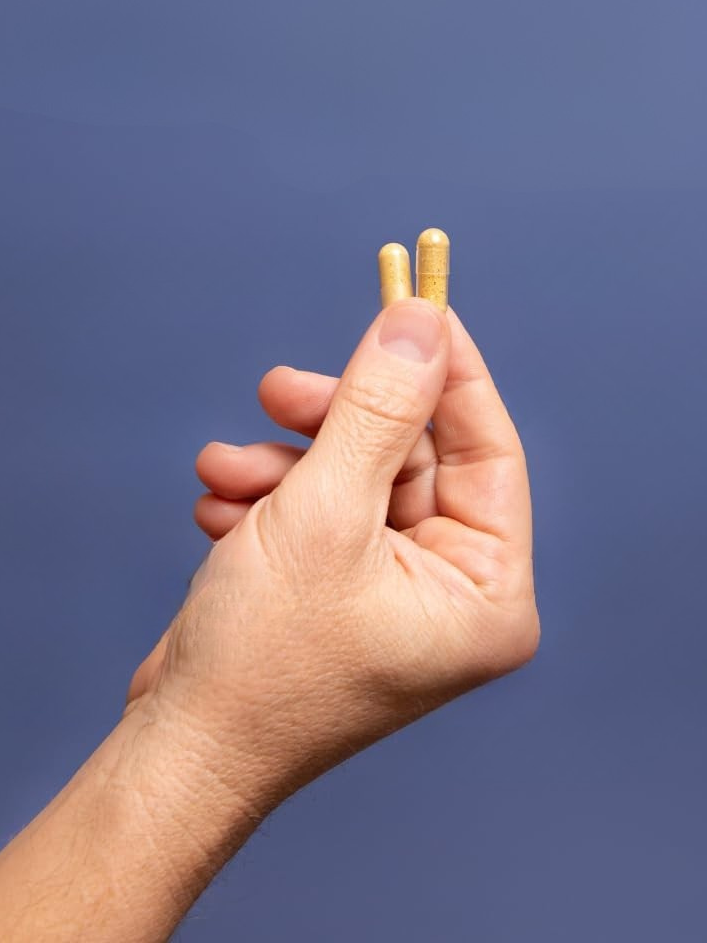 A hand holding two yellow capsules upright against a plain blue background.