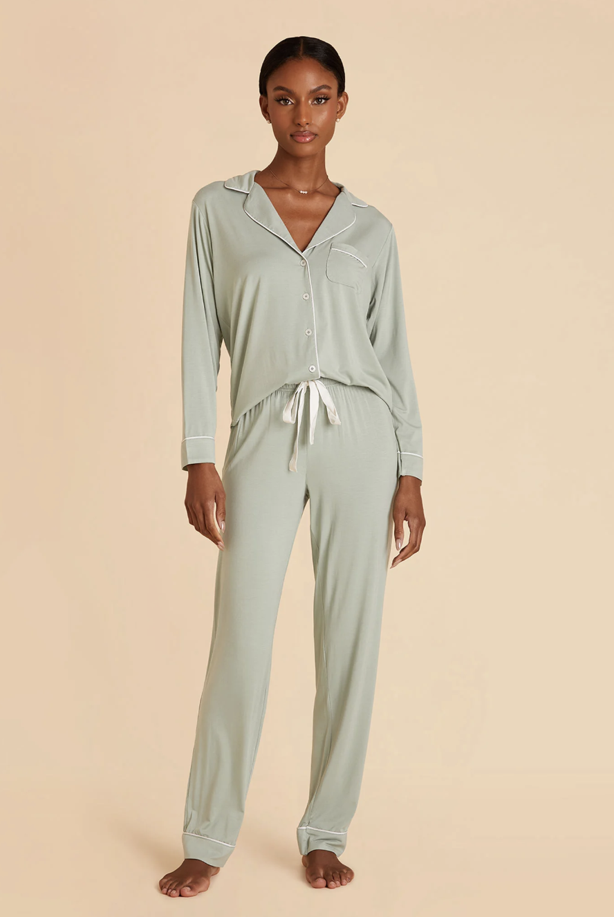 A woman stands barefoot wearing light sage green long-sleeve button-up pajamas with white trim and a drawstring waist, against a beige background.
