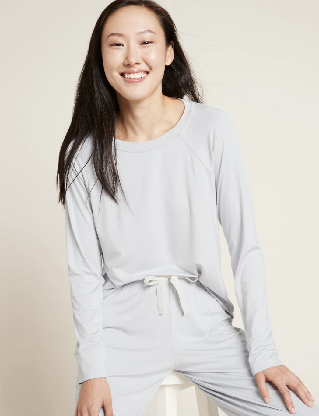 Woman in light gray long-sleeve top and matching drawstring pants sits on a stool and smiles at the camera against a neutral background.