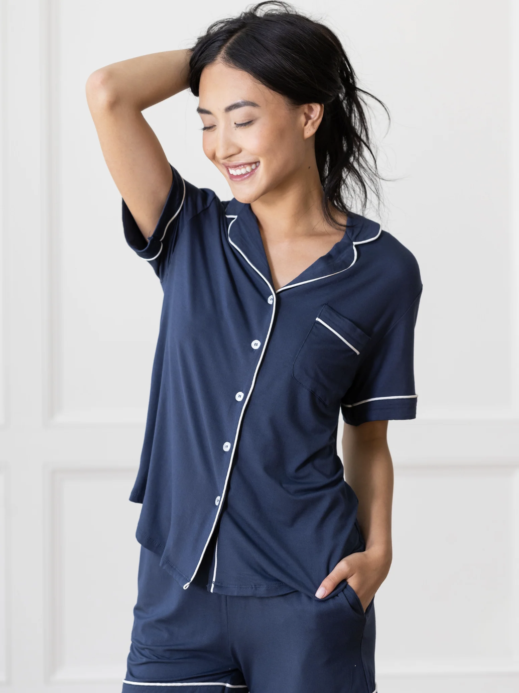 A person wearing navy blue short-sleeve pajamas with white piping stands with one hand in their pocket and the other hand touching their hair, smiling and looking to the side.