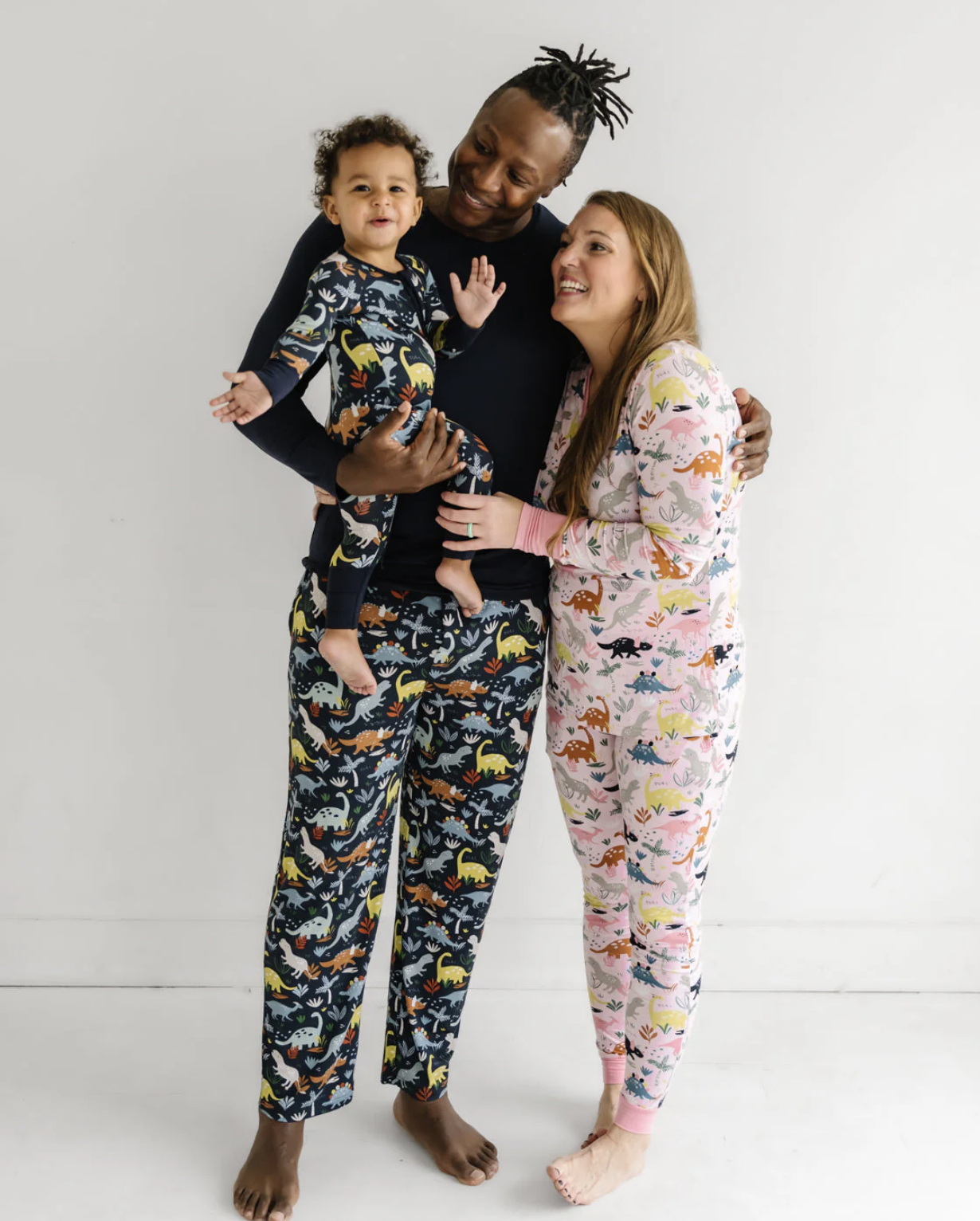 A family of three wearing dinosaur print pajamas stands together smiling against a plain white background.