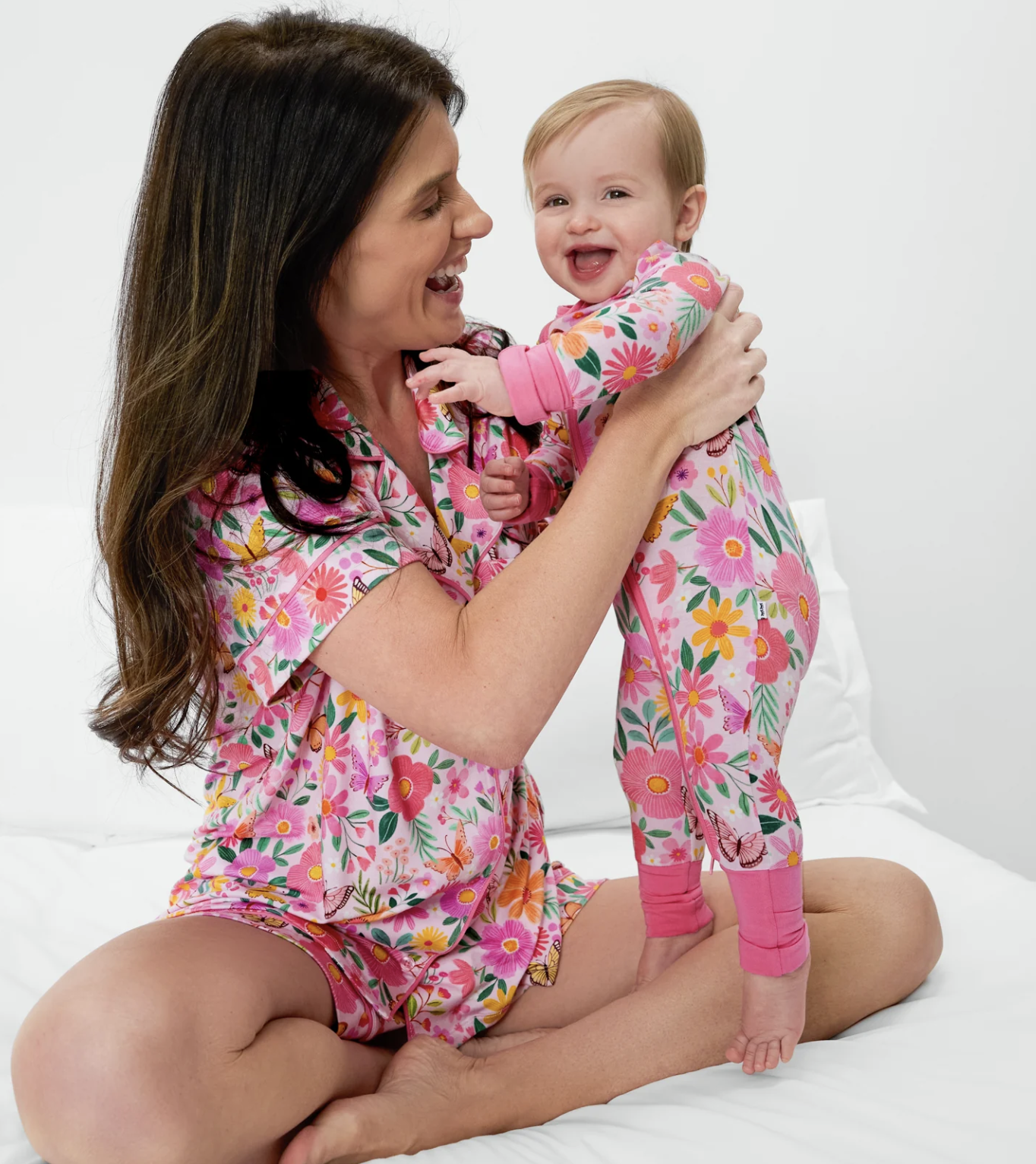 Woman sitting on a bed holding a smiling baby. Both are wearing matching pink floral pajamas.