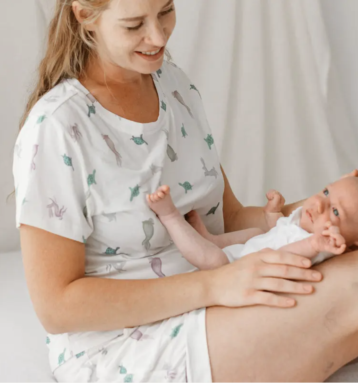 An adult sitting in bed holds a baby on her lap. Both are wearing light-colored clothing, and the adult's outfit has a cat pattern.