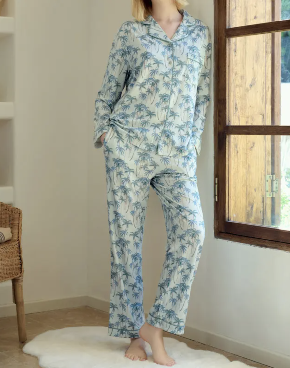 Person standing indoors near a window, wearing light-colored pajama set with a blue palm tree pattern, hands in pockets, barefoot on a white rug.