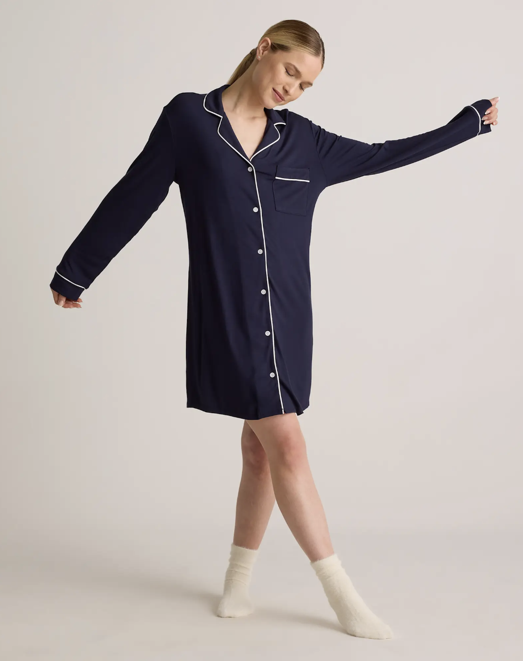 A woman wearing a long-sleeve, navy blue button-up nightshirt with white piping and white socks stands with arms extended against a plain background.