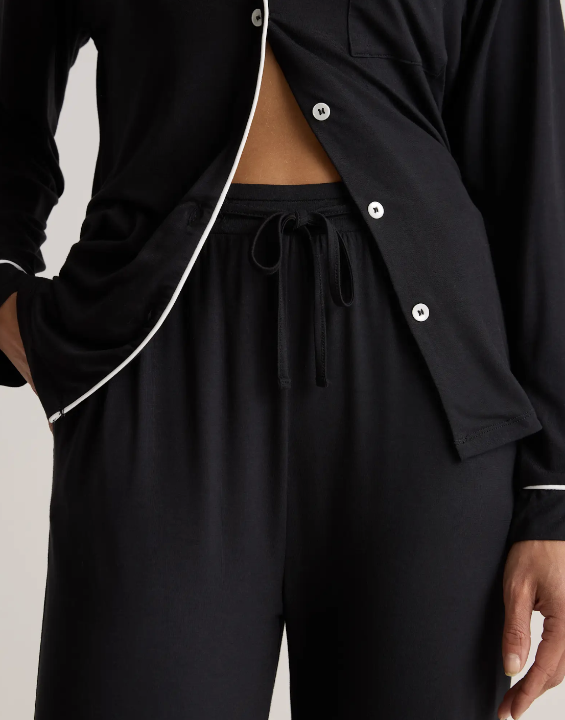 A person wearing black pajama pants and a matching black pajama shirt with white piping and buttons, one hand in their pocket.