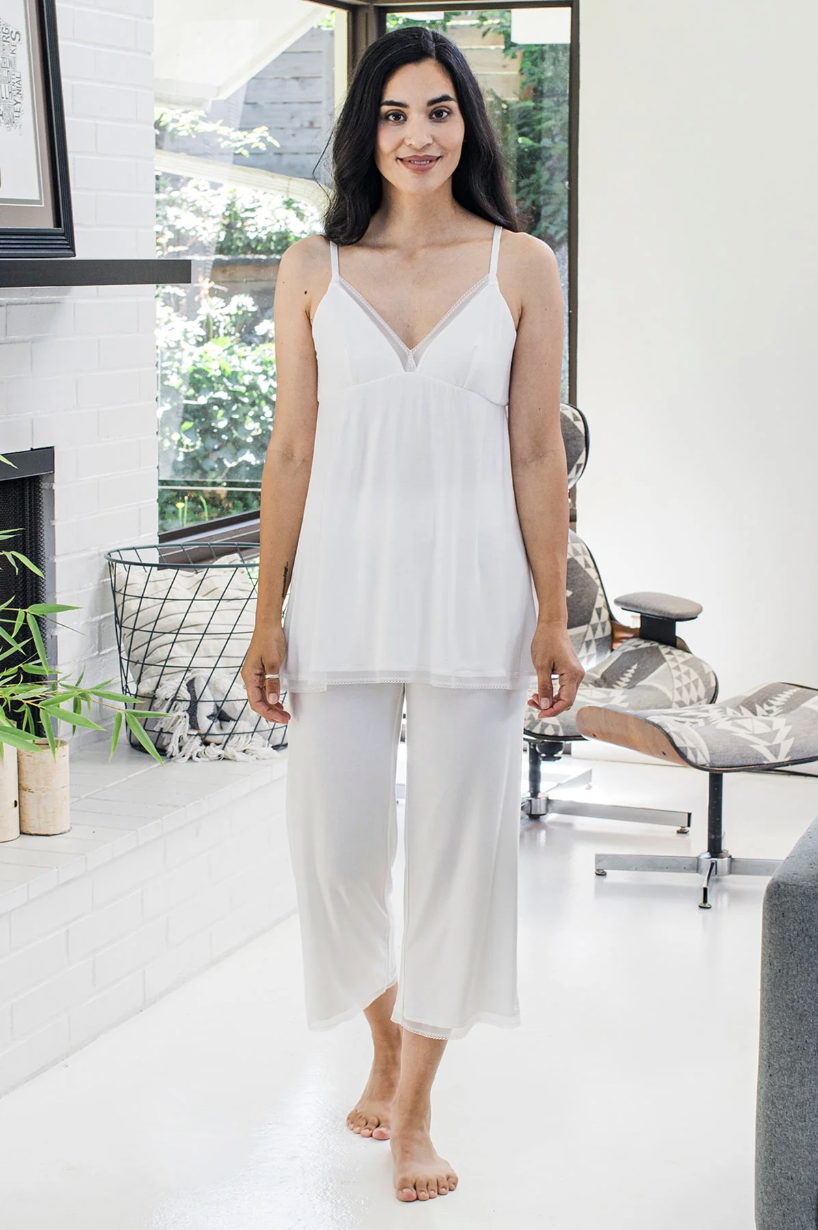 A woman stands barefoot indoors wearing a white sleeveless pajama set, with a relaxed fit camisole top and loose pants. She is smiling and facing the camera.