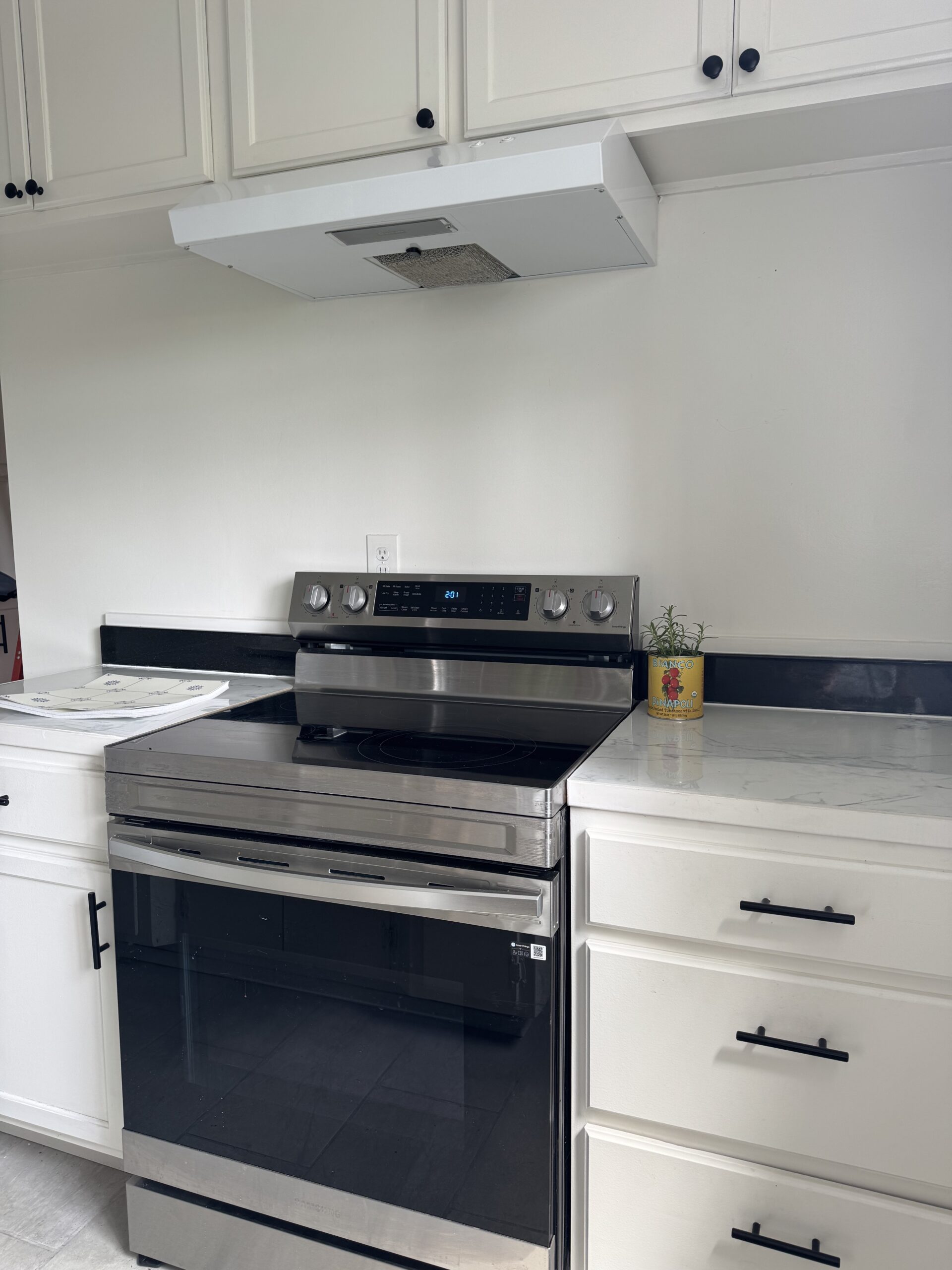 A modern kitchen with a stainless steel electric stove, white cabinets, black handles, marble countertops, and a potted herb on the right side.