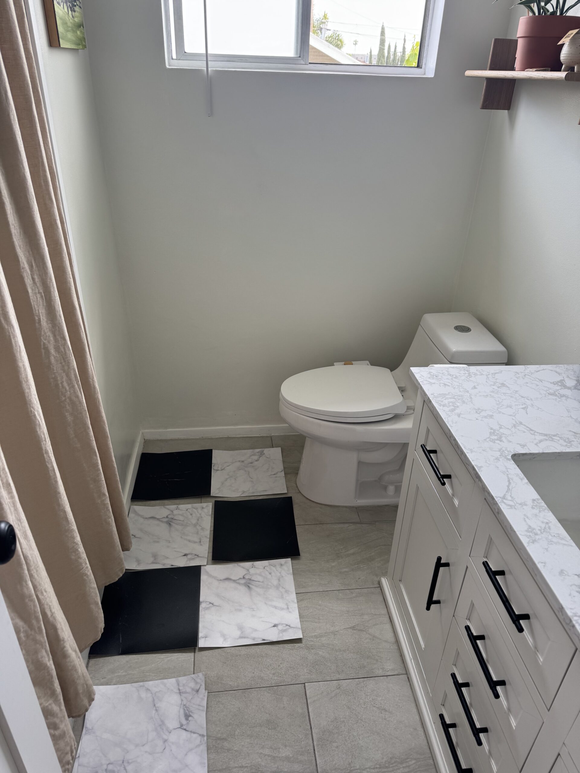 A bathroom with a toilet, a white vanity with drawers, a window, and a floor partially covered with black and white tiles placed on top of gray tiles.