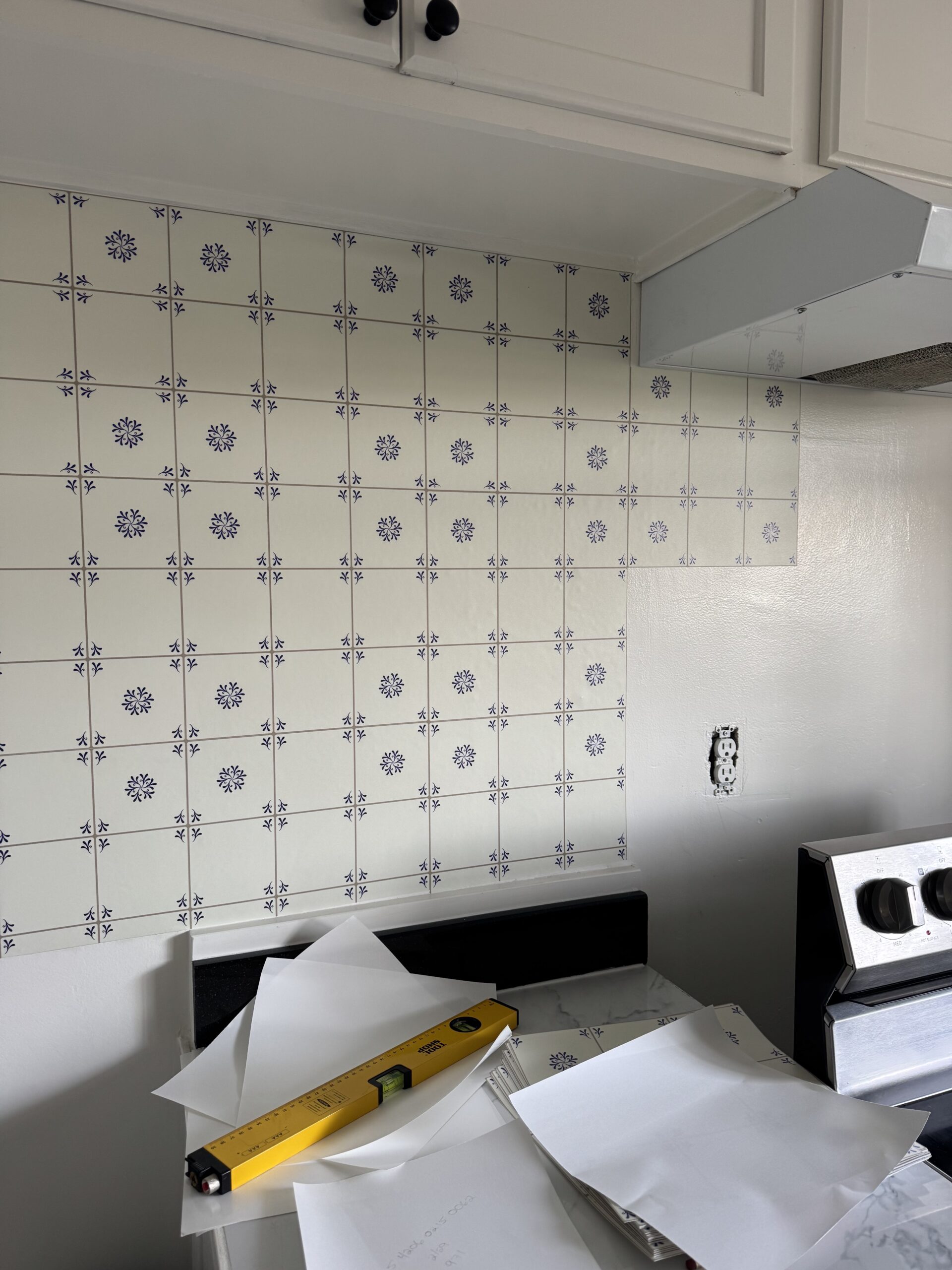 Partially installed patterned tile backsplash in a kitchen, with tiles, tools, and papers on the stove and nearby surfaces.