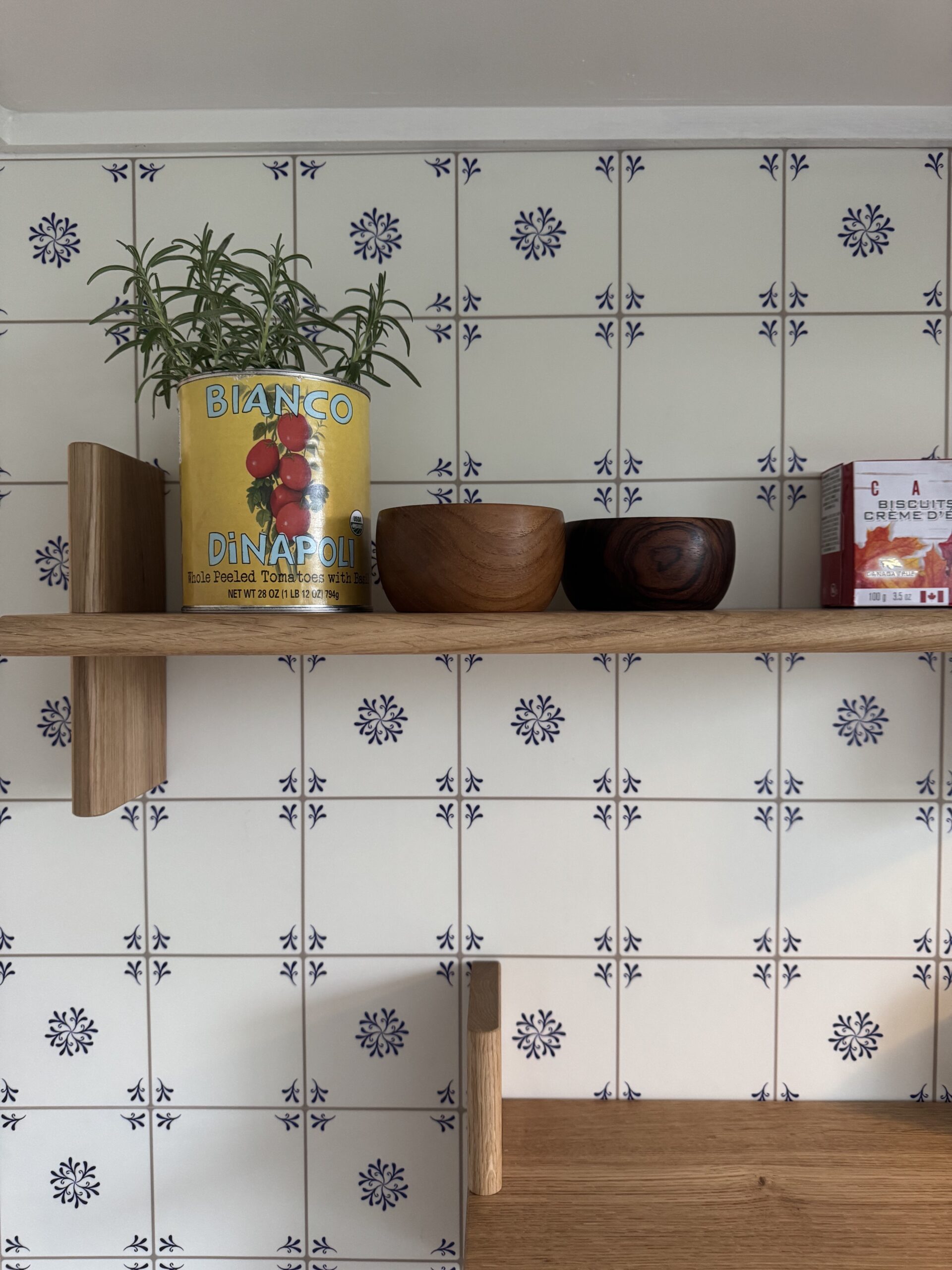 A wooden shelf holds a rosemary plant in a tomato can, two wooden bowls, and a box against a blue and white patterned tile wall.