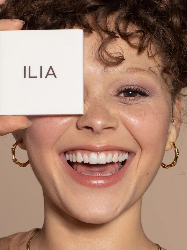 Smiling woman with curly hair holds a white ILIA cosmetics compact over one eye, wearing hoop earrings and natural makeup.