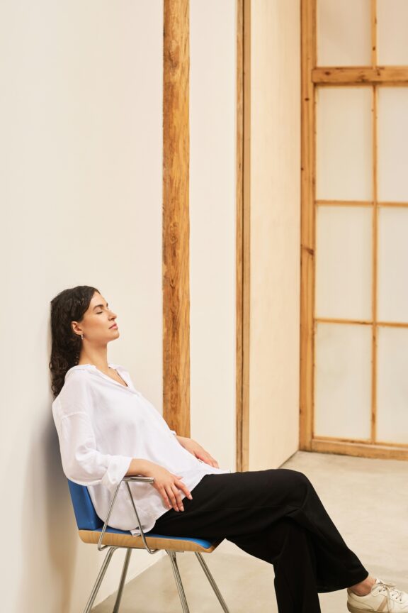 A woman in a white shirt and black pants sits on a blue chair, leaning back against a white wall in a minimal, modern room with wooden beams and a large window.