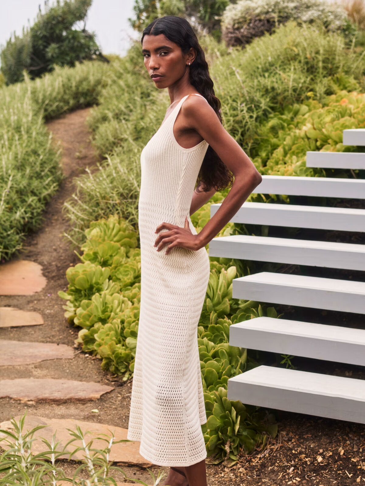A woman in a sleeveless, ankle-length beige knit dress stands outdoors next to gray stairs, surrounded by greenery and plants.