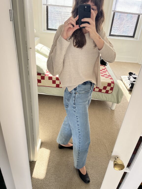 Person taking a mirror selfie in a bright bedroom, wearing a light sweater, blue jeans, and black shoes. A bed with a red and white quilt is visible in the background.